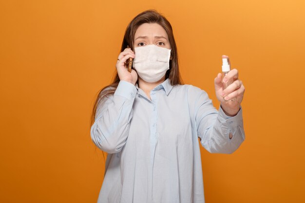 Portret van een jonge vrouw op gele muur praten over de telefoon, paniek, spray antiseptische in de hand