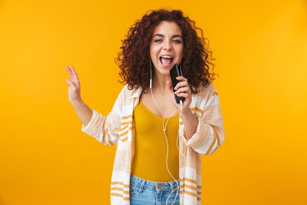 Portret van een jonge vrouw van 20 jaar met een koptelefoon die zingt en luistert naar muziek op een smartphone, geïsoleerd op geel