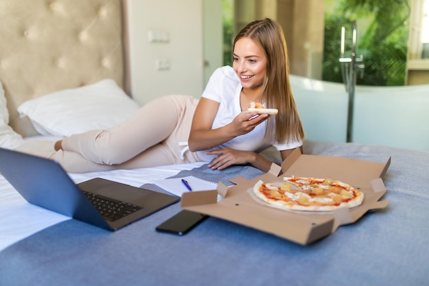 Portret van een jonge vrouw zittend op een bed met een doos pizza, een stuk pizza bijtend