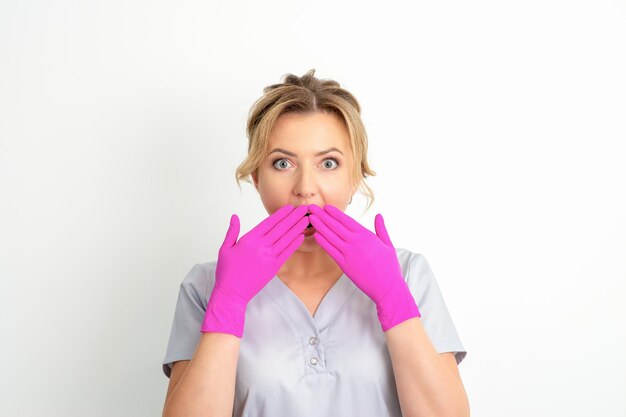 Portret van een jonge vrouwelijke blanke arts of verpleegster is geschokt door haar mond te bedekken met haar roze gehandschoende handen tegen een witte achtergrond.