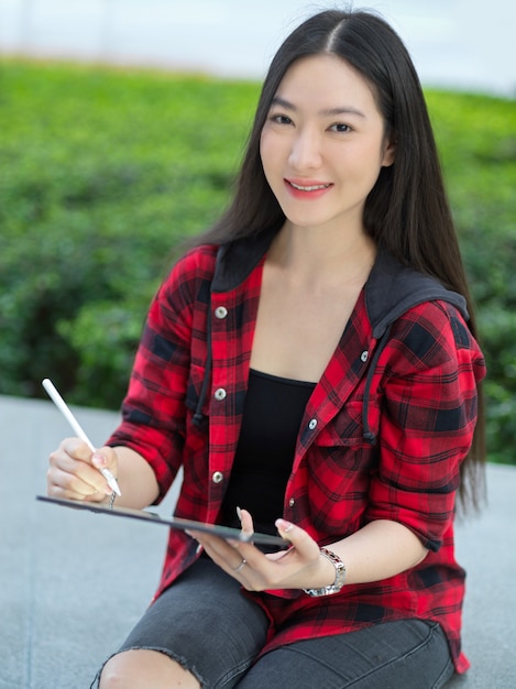Portret van een jonge vrouwelijke student die digitale tablet gebruikt met een styluspen die in het park zit