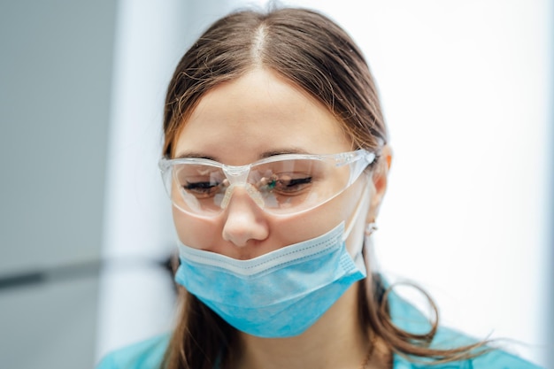 Portret van een jonge vrouwelijke tandarts met een chirurgisch masker en een veiligheidsbril Schoonheidsgezicht van een arts of verpleegster die een beschermend masker en een bril draagt in de kliniek Close-up