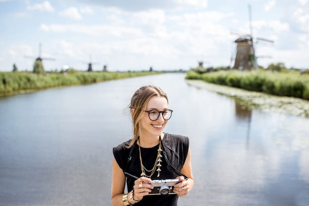 Portret van een jonge vrouwelijke toerist die met een fotocamera op de prachtige landschapsachtergrond staat met oude windmolens in Nederland