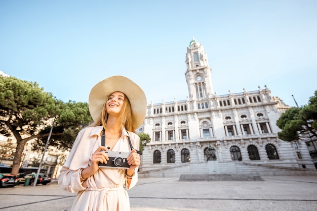 Portret van een jonge vrouwentoerist in zonnehoed die zich met fotocamera voor het stadhuisgebouw bevindt tijdens het ochtendlicht in Porto, Portugal