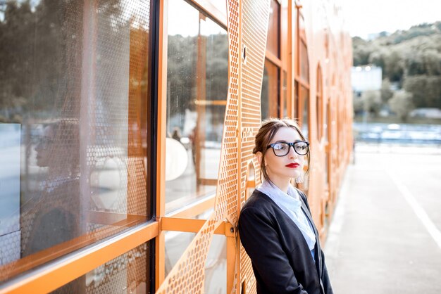 Portret van een jonge zakenvrouw in het pak die buiten op de moderne kantoormuur staat