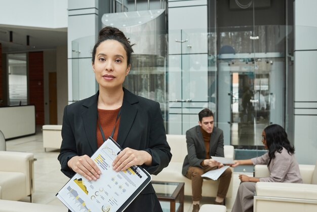 Portret van een jonge zakenvrouw met een verslag dat naar de camera kijkt met haar collega's op de achtergrond