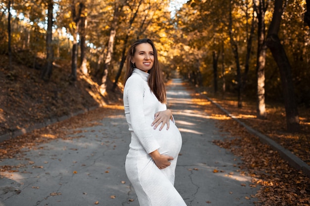 Portret van een jonge zwangere gelukkige vrouw die de buik aanraakt met handen die een witte jurk dragen op de achtergrond van een gouden herfstpark