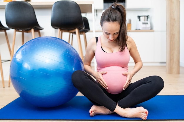 Portret van een jonge zwangere vrouw die op een oefenmat zit en haar buik aanraakt in de woonkamer in haar huis