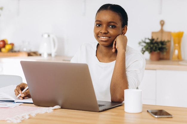 Portret van een jonge zwarte vrouw die in de keuken zit en thuis op een laptop werkt.