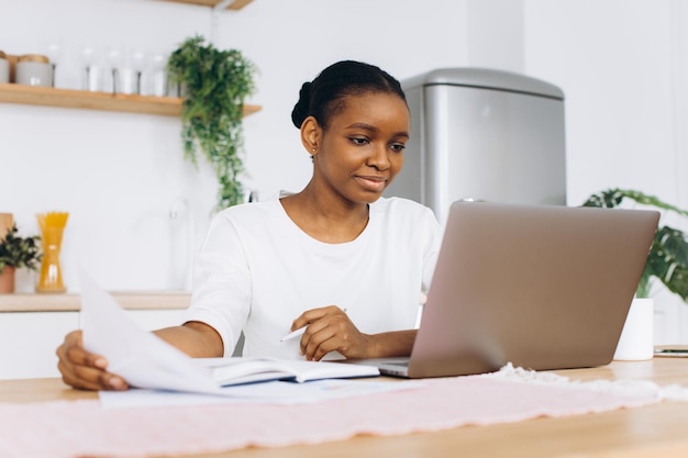 Portret van een jonge zwarte vrouw die in de keuken zit en thuis op een laptop werkt