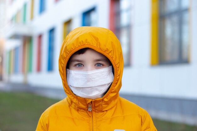 Portret van een jongen in een gele jas en capuchon in een witte medische masker op straat