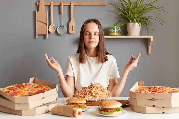 Portret van een kalme, ontspannen, dankbare vrouw met bruin haar, gekleed in een wit casual T-shirt, zittend aan tafel in de keuken die yoga beoefent of mediteert voordat ze een smakelijk diner eet met pizza pasta hamburger