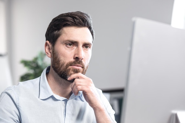 Portret van een kantoormedewerker Nadenkend serieus met een baard Zittend aan een bureau op kantoor op de computer in een wit overhemd