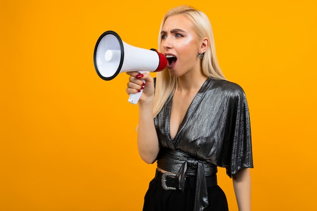 Portret van een Kaukasisch blond meisje in een grafietblouse die nieuws in een megafoon op een gele studioachtergrond spreekt