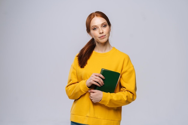 Portret van een kaukasische jonge vrouw die een boek vasthoudt en naar de camera kijkt op een geïsoleerde grijze achtergrond Mooie roodharige dame die vrijetijdskleding draagt en emotioneel gezichtsuitdrukkingen toont