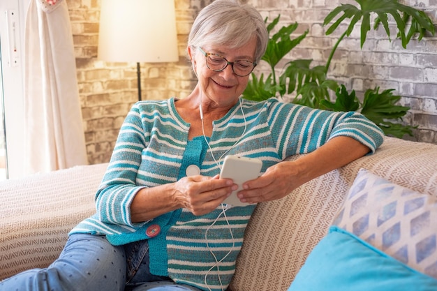 Portret van een kaukasische senior vrouw met een koptelefoon die thuis op de bank zit, muziek luistert of video kijkt op een mobiele telefoon Bakstenen muur achtergrond