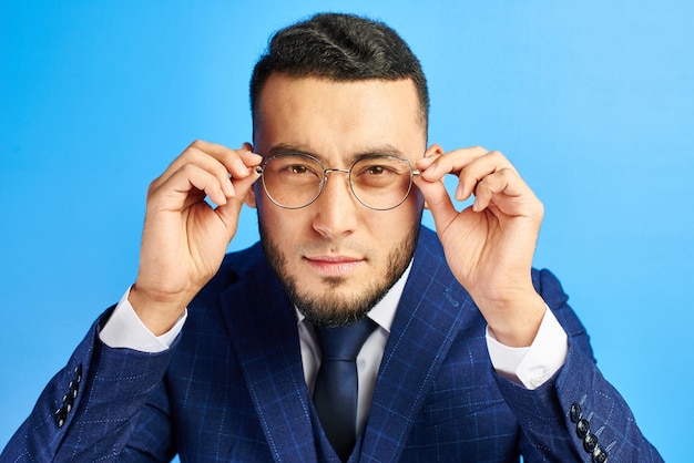 Portret van een Kazachse Aziatische zakenman in een pak en bril, nieuwsgierig naar je kijkend, loensend, proberend dichterbij te kijken op een blauwe achtergrond in de studio