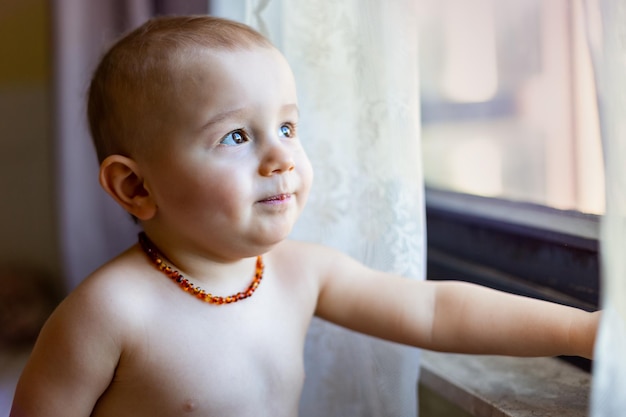 Portret van een kind dat 's ochtends naar het raam kijkt Een peuterjongen die een amberkleurige ketting draagt, pijnloos tandjes krijgen, een natuurlijke pijnstiller