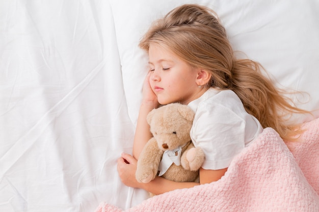 portret van een klein blond meisje dat in een bed slaapt met een teddybeer onder een roze deken