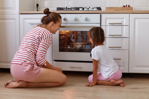 Portret van een klein donkerharige jongen kijken met haar moeder in de oven koekjes bakken in de oven binnenshuis familie dragen casual kleding zittend op de vloer in de keuken