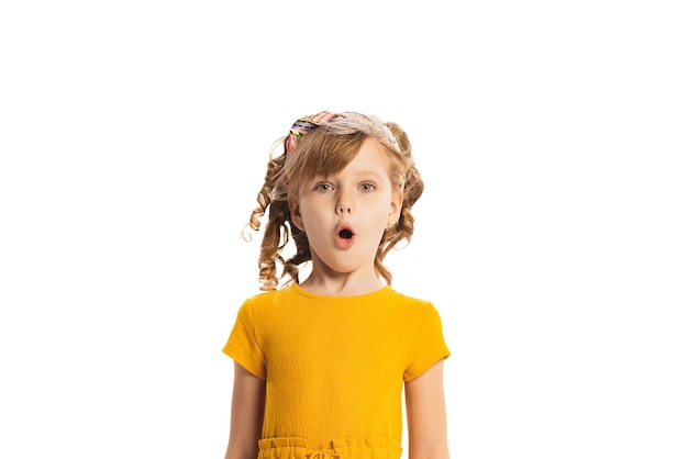 Portret van een klein emotioneel kindmeisje in een gele jurk die zich voordeed en gezichten maakt geïsoleerd over een witte studio