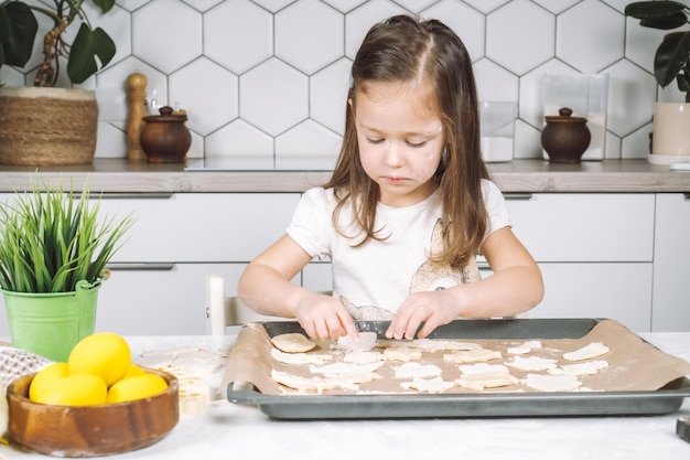 Portret van een klein leergierig meisje dat in de stoel zit en in de keuken verschillende deegkoekjes in de vorm van Pasen maakt