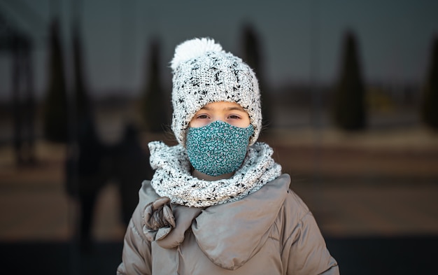 Portret van een klein meisje dat een hoed en een masker draagt tijdens de coronaviruspandemie.