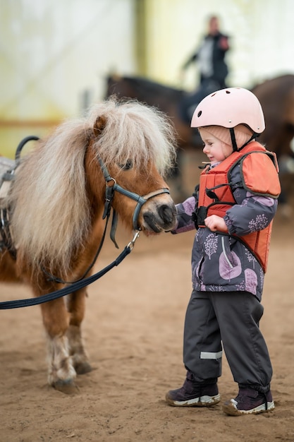 Portret van een klein meisje in een beschermend jasje en helm met haar bruine pony voor rijles