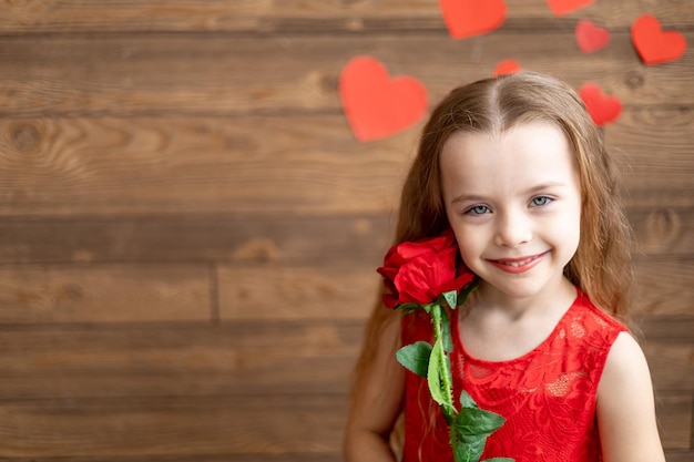 Portret van een klein meisje in een rode jurk met een rode roos op een donkerbruine houten achtergrond en zoet glimlachend het concept van Valentijnsdag een lege ruimte voor tekst