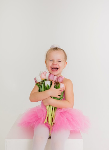 Portret van een klein meisje in een roze jurk met een tutu rokje zittend met een boeket tulpen op een witte muur