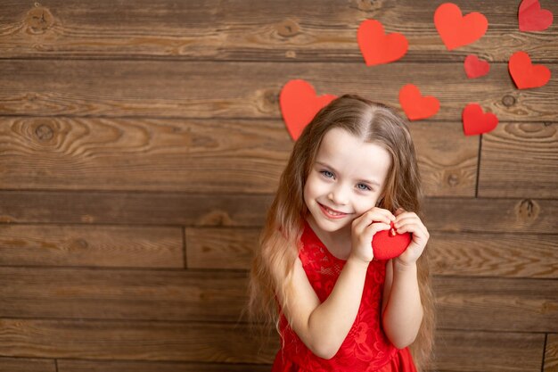 Portret van een klein meisje kind in een rode jurk met een rood hart speelgoed op een donkerbruine houten achtergrond en lief glimlachend het concept van Valentijnsdag een lege ruimte voor tekst