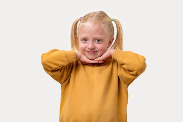 Portret van een klein meisje met blond haar in een gele sweatshirt die haar handen vasthoudt aan haar kin