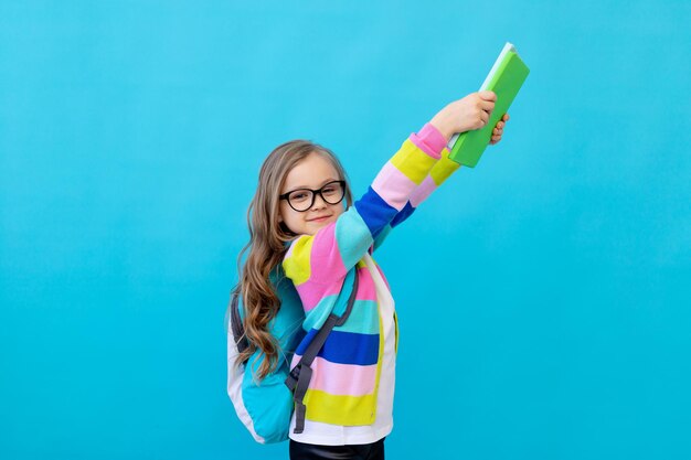 Portret van een klein meisje met een bril in een gestreept jasje met notitieboekjes en schoolboeken in haar handen en een rugzak. Het concept van onderwijs. Fotostudio, blauwe achtergrond, plaats voor tekst