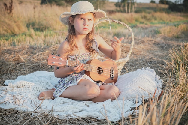 Portret van een klein meisje met een hoed zittend op een deken in een droog hooiveld met een picknick die ukelele leert spelen