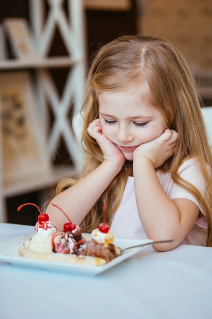 Portret van een klein mooi meisje, zittend in een café aan de tafel te denken met een bolletje ijs met fruit. gelukkig kind