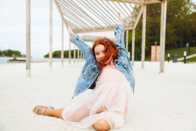 Portret van een klein roodharig meisje in een jurk zittend op het zand van het strand, zonnige zomerdag