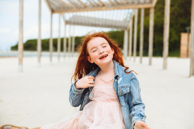 Portret van een klein roodharig meisje in een jurk zittend op het zand van het strand, zonnige zomerdag