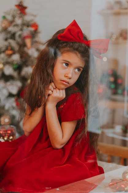 Foto portret van een klein schattig meisje in een rode jurk met een strik op haar hoofd door glas close-up