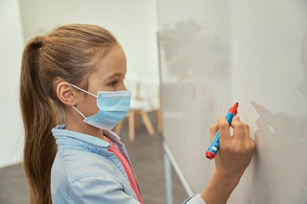 Portret van een klein schoolmeisje dat een beschermend masker draagt tijdens het schrijven van een pandemie van het coronavirus