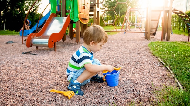 Portret van een kleine 3 jaar oude peuterjongen die op de speelplaats zit en zand graaft met een plastic speelgoedschep