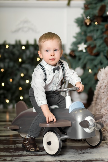 Portret van een kleine jongenszitting op een uitstekend stuk speelgoed vliegtuig dichtbij een Kerstboom. Kerst versiering. De jongen verheugt zich op zijn kerstcadeau. Vrolijk kerstfeest en een gelukkig nieuwjaar