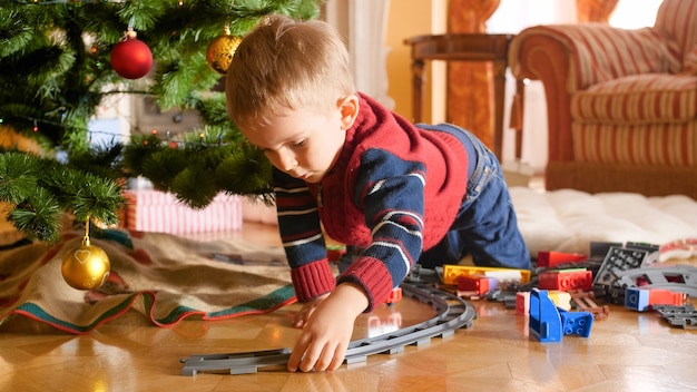 Portret van een kleine peuterjongen die een spoorweg bouwt en speelt met een speelgoedtrein onder de kerstboom