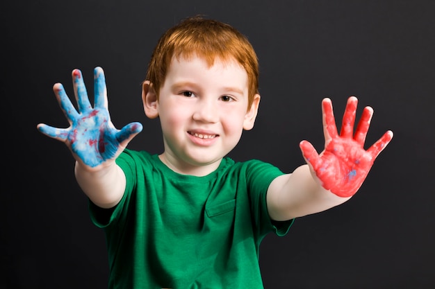 Portret van een kleine roodharige jongen tekent op papier met verf in verschillende kleuren die de jongen met zijn handen tekent