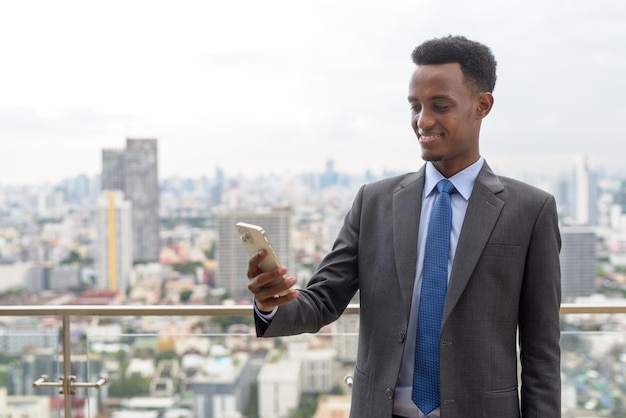 Portret van een knappe Afrikaanse zakenman met een pak en stropdas op het dak in de stad tijdens het gebruik van een mobiele telefoon