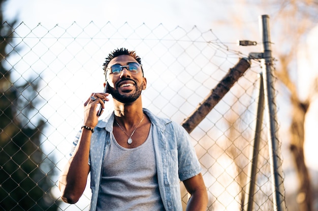 Portret van een knappe Afro-Amerikaanse man die zijn smartphone op straat gebruikt en glimlachend op een hek leunt