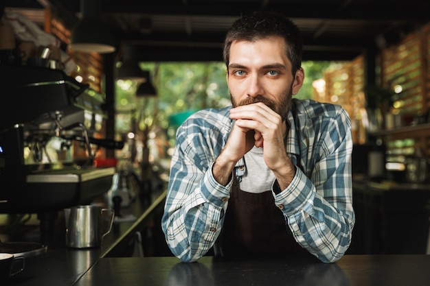 Portret van een knappe barista-man met een schort die lacht terwijl hij in een straatcafé of een koffiehuis buiten werkt