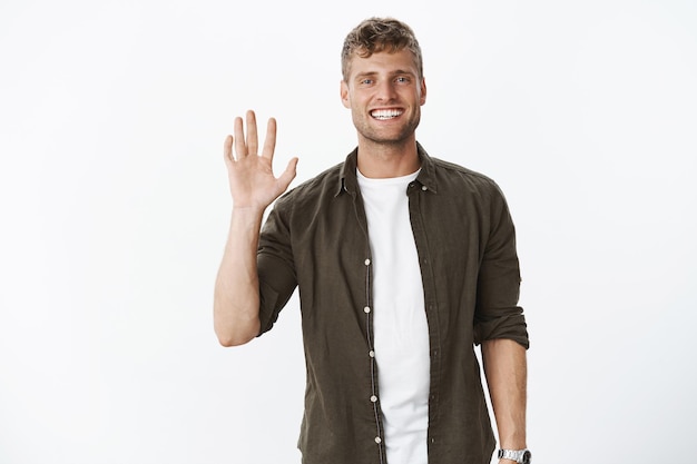 Portret van een knappe, charismatische en mannelijke man met blauwe ogen en een witte glimlach die met opgeheven hand zwaait en ons begroet met een aangename vriendelijke blik over de grijze muur