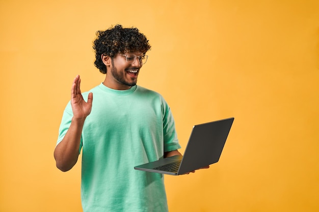 Foto portret van een knappe gekrulde indiase man in een turquoise t-shirt en bril die een laptop vasthoudt en hallo zegt terwijl hij tegen een gele achtergrond staat