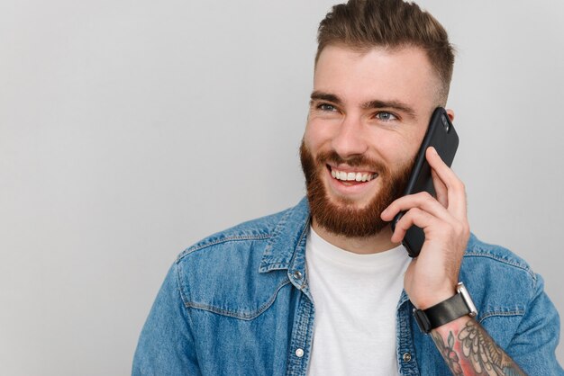 Portret van een knappe glimlachende jonge man met vrijetijdskleding die geïsoleerd staat over een grijze muur, pratend op mobiele telefoon