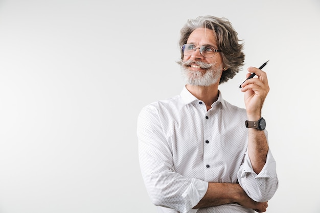 Portret van een knappe glimlachende volwassen zakenman gekleed in formele kleding die geïsoleerd over een witte muur staat, een pen vasthoudt en wegkijkt